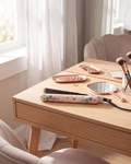 A floral-patterned hair straightener and matching rose gold makeup trays and mirror set on a light wood vanity table in a bright room.