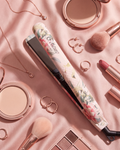 A floral-patterned hair straightener styled in a flat lay with pink makeup items, jewelry, and brushes on a pink satin background.
