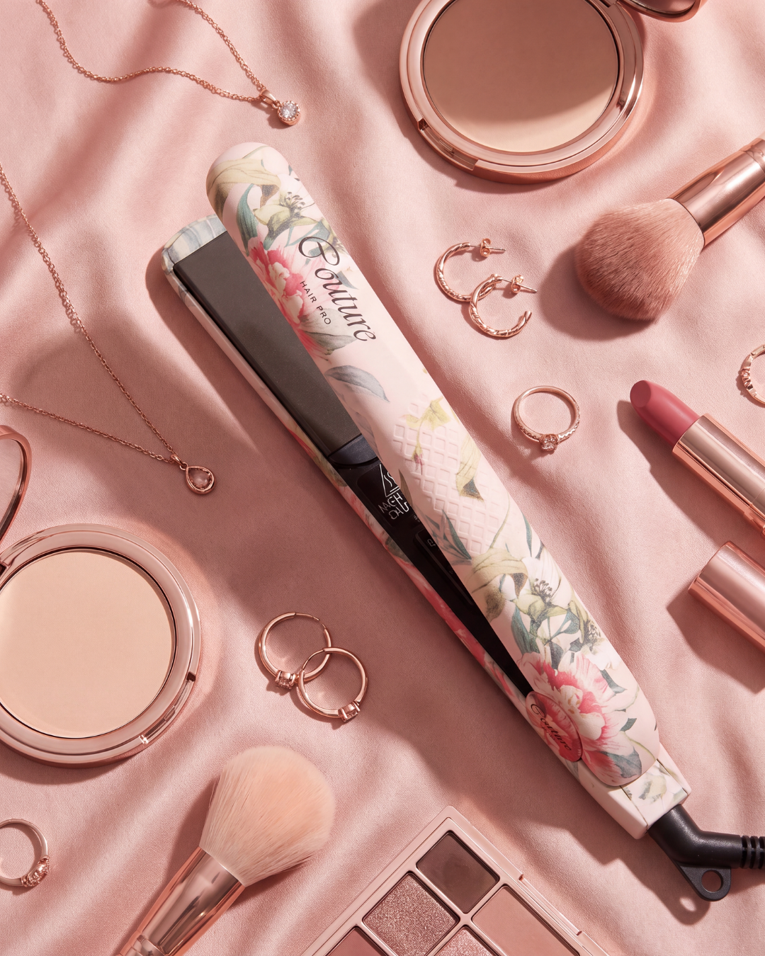 A floral-patterned hair straightener styled in a flat lay with pink makeup items, jewelry, and brushes on a pink satin background.