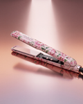 A hair straightener with a pink floral pattern, shown open on a reflective surface against a warm pink background. The brand name 'couture' is visible on the plates.