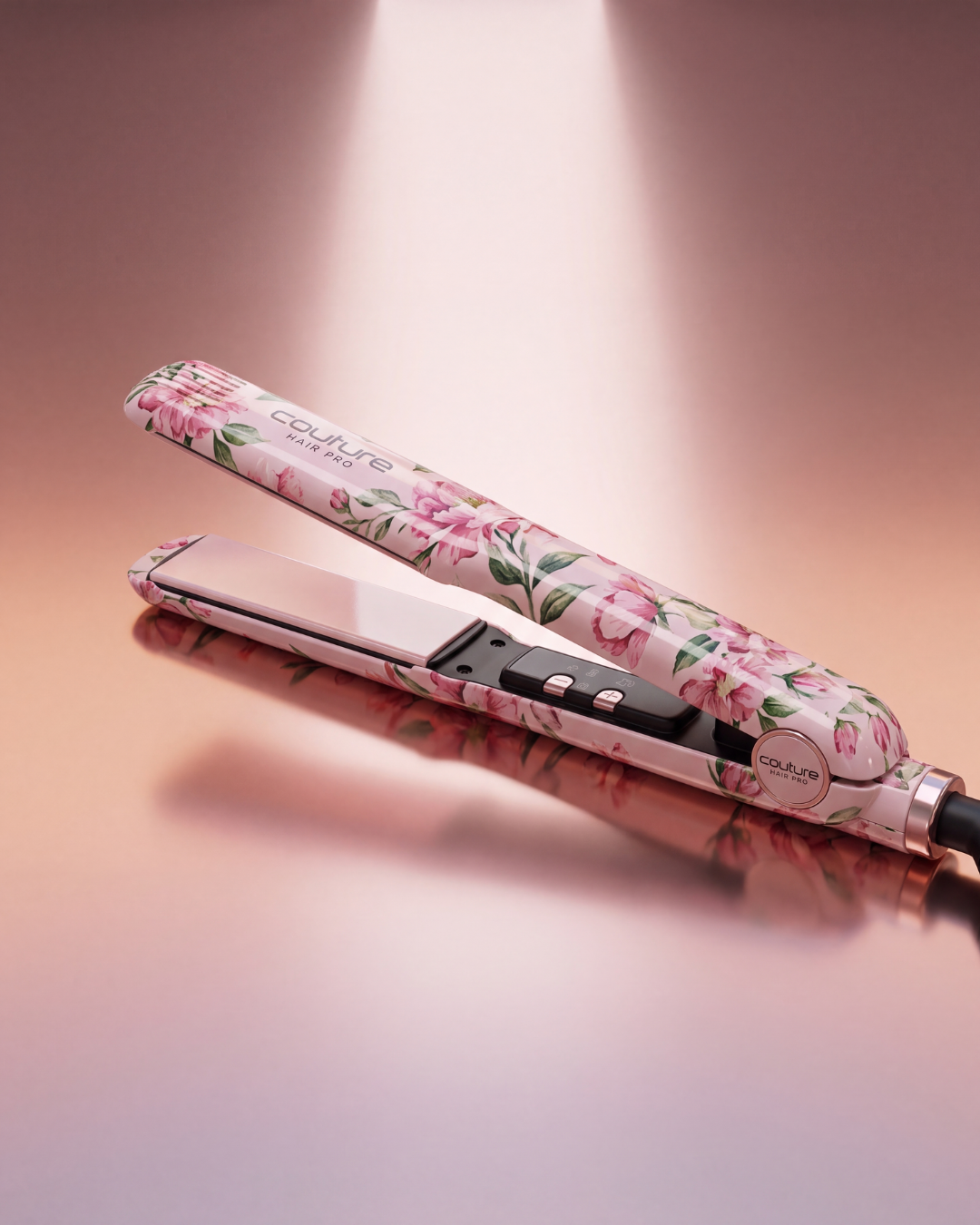 A hair straightener with a pink floral pattern, shown open on a reflective surface against a warm pink background. The brand name 'couture' is visible on the plates.