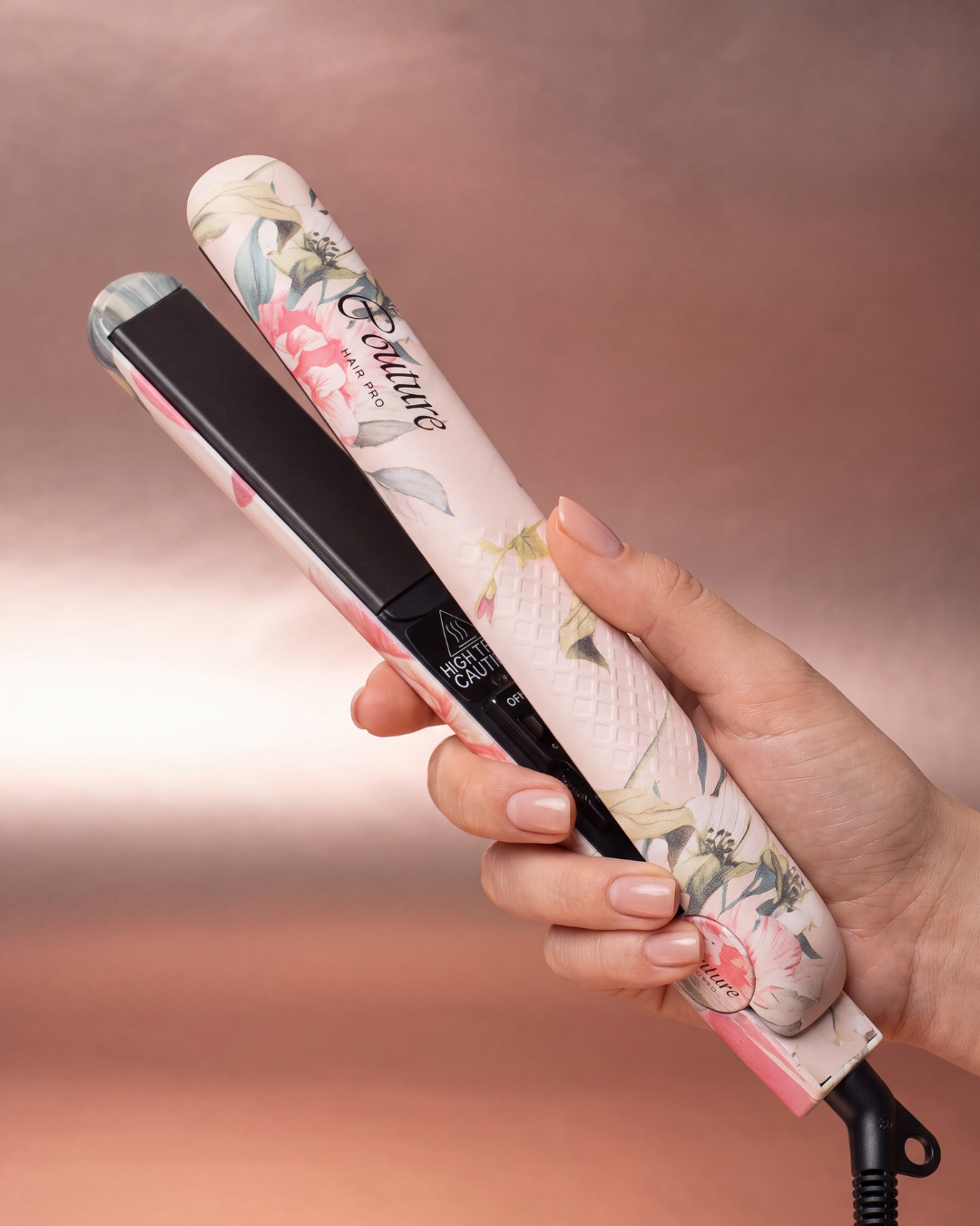 A hand holding a pink floral hair straightener with black plates against a soft pink background.